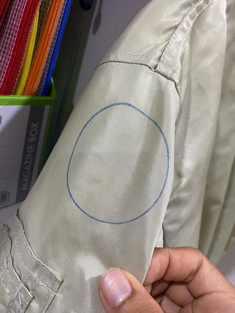 May include: Close-up of a light green jacket sleeve. A blue circle is drawn on the sleeve. The jacket has a pocket and a seam detail. The sleeve is being held by a person's hand.