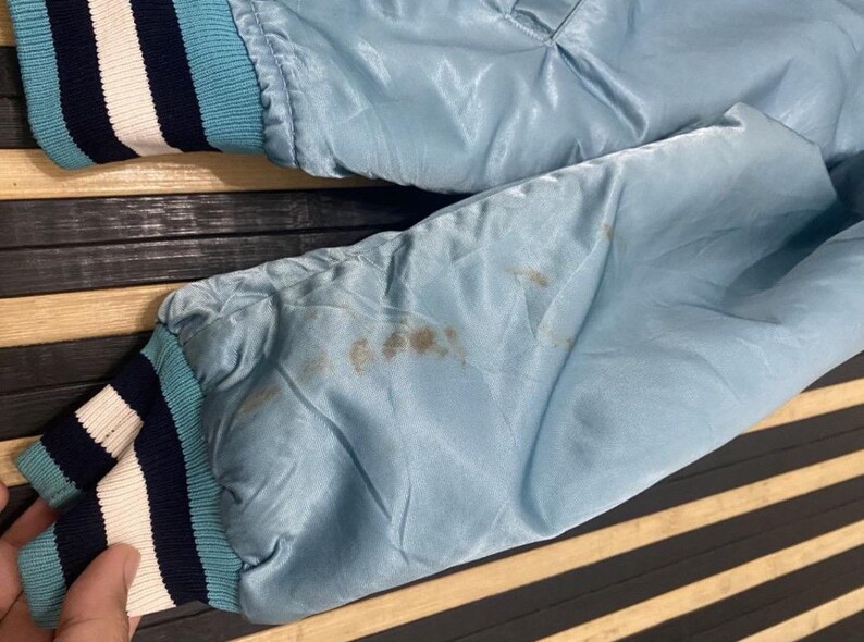 Puede incluir: Chaqueta de sat&eacute;n azul claro con cuello y pu&ntilde;os de canal&eacute;. Los pu&ntilde;os presentan rayas azul marino, blancas y azul claro. La chaqueta tiene manchas en la manga.