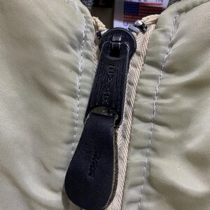 May include: Close-up of a vintage olive green bomber jacket zipper. The black zipper pull is leather with the brand name "Emar" embossed. The jacket has a gray collar and a tag with the text "PROPERTY U.S. AIR FORCE" and a small American flag.