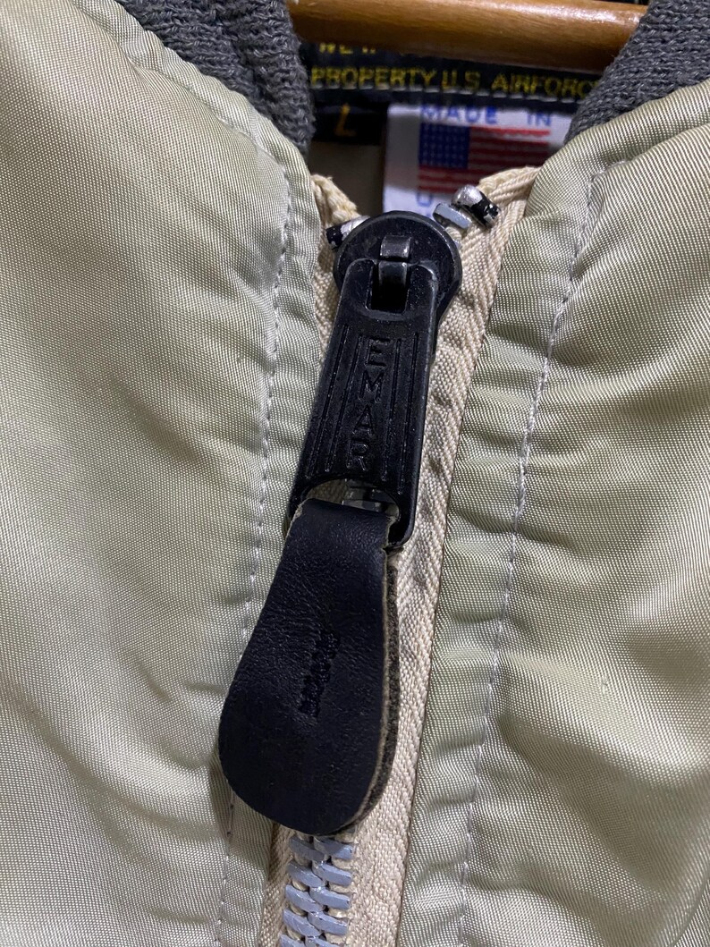 May include: Close-up of a vintage olive green bomber jacket zipper. The black zipper pull is leather with the brand name "Emar" embossed. The jacket has a gray collar and a tag with the text "PROPERTY U.S. AIR FORCE" and a small American flag.