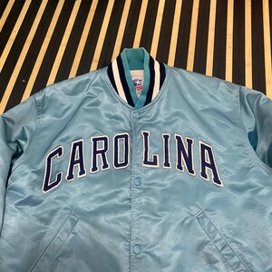 Puede incluir: Chaqueta de sat&eacute;n azul claro con la palabra "CAROLINA" en letras azul marino y blanco en el pecho. La chaqueta tiene un cuello a rayas verde azulado, azul marino y blanco y una parte delantera abotonada. Una chaqueta vintage de tem&aacute;tica deportiva.