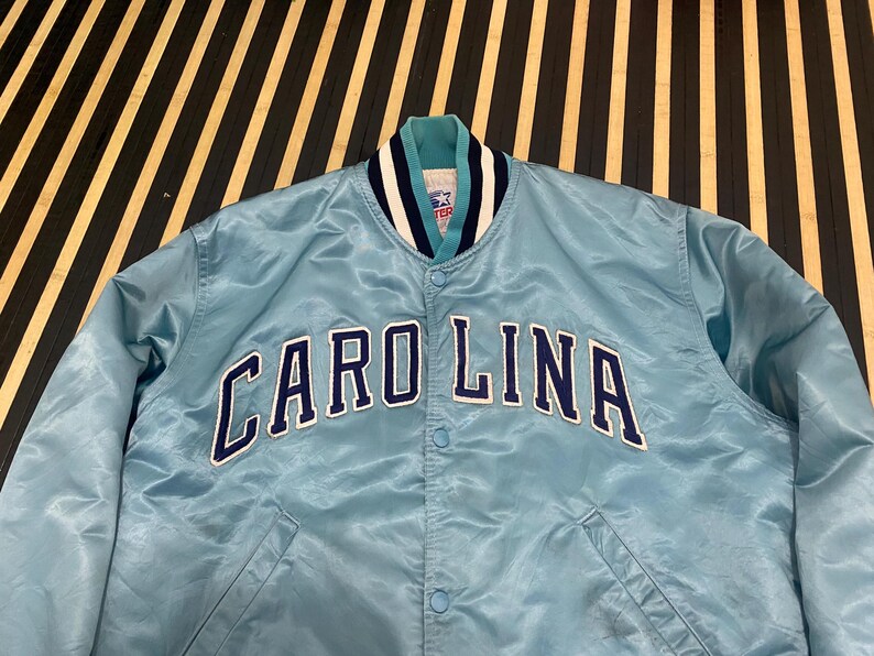 Puede incluir: Chaqueta de sat&eacute;n azul claro con la palabra "CAROLINA" en letras azul marino y blanco en el pecho. La chaqueta tiene un cuello a rayas verde azulado, azul marino y blanco y una parte delantera abotonada. Una chaqueta vintage de tem&aacute;tica deportiva.