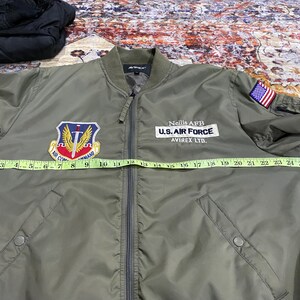 May include: An olive green bomber jacket with a front zipper, featuring an Air Command patch, a Nellis AFB U.S. Air Force Avirex Ltd. patch, and a small American flag patch on the sleeve. A yellow measuring tape shows a width of 27 inches.
