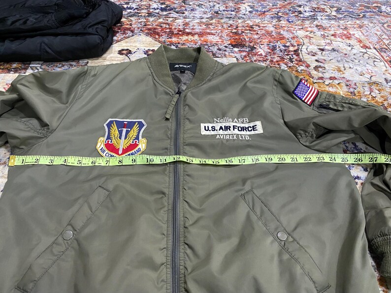 May include: An olive green bomber jacket with a front zipper, featuring an Air Command patch, a Nellis AFB U.S. Air Force Avirex Ltd. patch, and a small American flag patch on the sleeve. A yellow measuring tape shows a width of 27 inches.