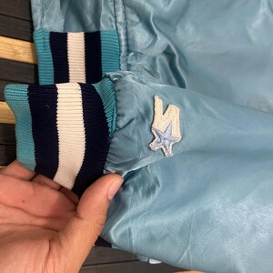 Puede incluir: Chaqueta de sat&eacute;n azul claro con pu&ntilde;os a rayas en azul marino, blanco y turquesa. Un peque&ntilde;o parche con dise&ntilde;o de estrella es visible en la manga. Chaqueta de estilo vintage.