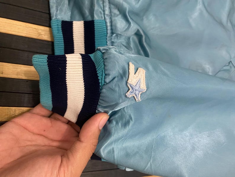 Puede incluir: Chaqueta de sat&eacute;n azul claro con pu&ntilde;os a rayas en azul marino, blanco y turquesa. Un peque&ntilde;o parche con dise&ntilde;o de estrella es visible en la manga. Chaqueta de estilo vintage.