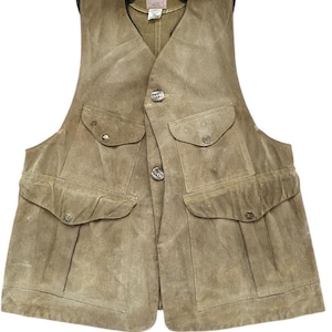 Peut inclure: Un gilet en daim beige avec quatre poches avant et une fermeture boutonn&eacute;e. Le gilet a un col vert et une &eacute;tiquette avec la marque "Filson". Le gilet est con&ccedil;u pour les activit&eacute;s de plein air et la chasse.