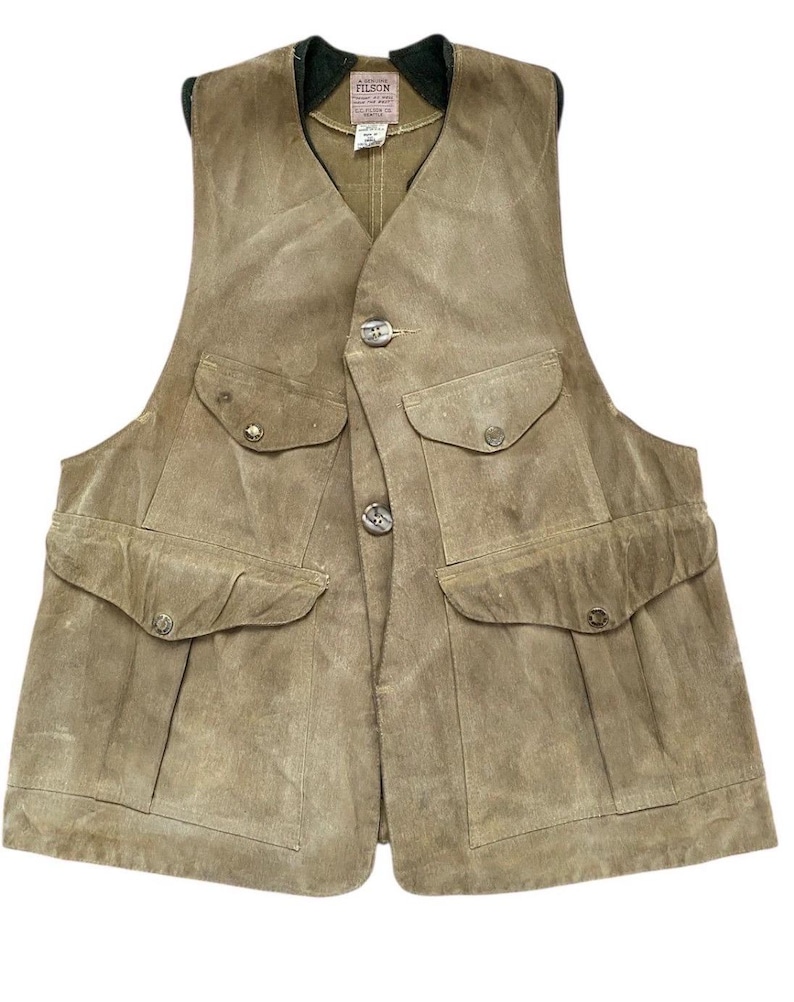 Peut inclure: Un gilet en daim beige avec quatre poches avant et une fermeture boutonn&eacute;e. Le gilet a un col vert et une &eacute;tiquette avec la marque "Filson". Le gilet est con&ccedil;u pour les activit&eacute;s de plein air et la chasse.
