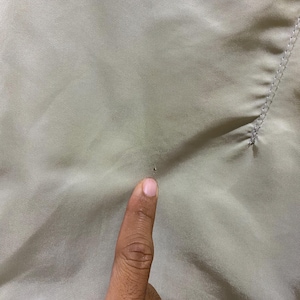May include: Close-up of a light olive-colored fabric. The fabric has a smooth texture with subtle diagonal lines. A finger is pointing at a small dark spot on the fabric.
