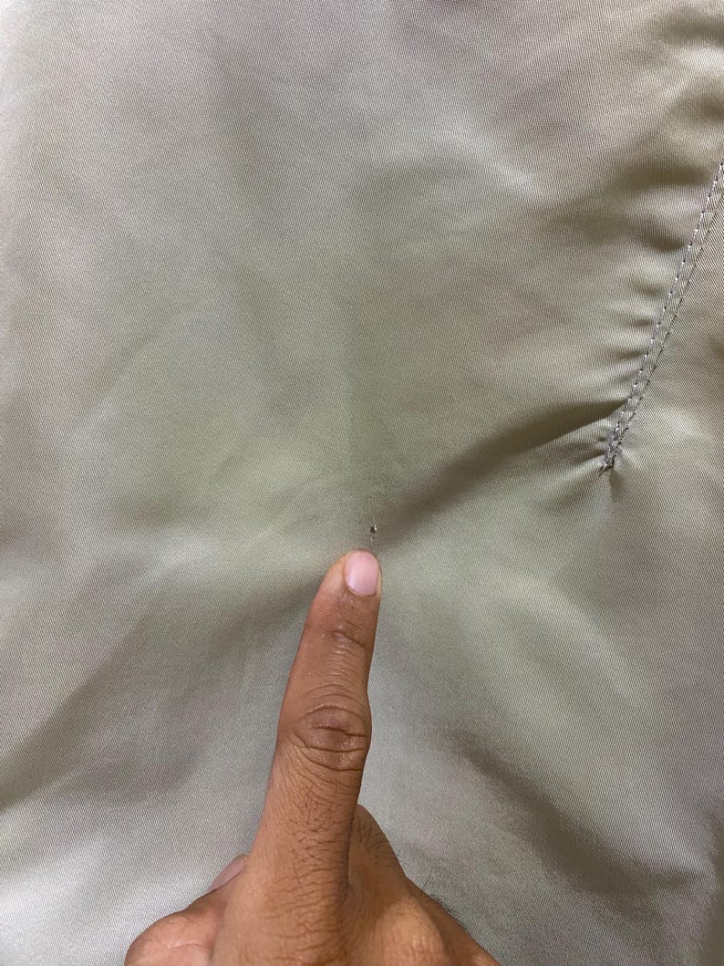May include: Close-up of a light olive-colored fabric. The fabric has a smooth texture with subtle diagonal lines. A finger is pointing at a small dark spot on the fabric.
