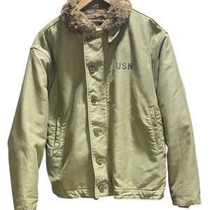 May include: An olive green jacket with a faux fur collar and button closure. The jacket has two front pockets and the letters "USN" printed on the chest. The jacket is hanging on a wooden hanger.
