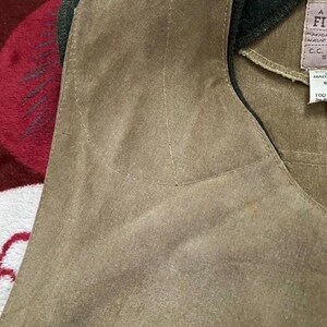 Peut inclure: Gros plan d'un gilet en daim beige avec un col vert fonc&eacute;. Le gilet a un d&eacute;tail de poche et des coutures visibles. Une &eacute;tiquette avec du texte est visible sur le c&ocirc;t&eacute; droit du gilet.