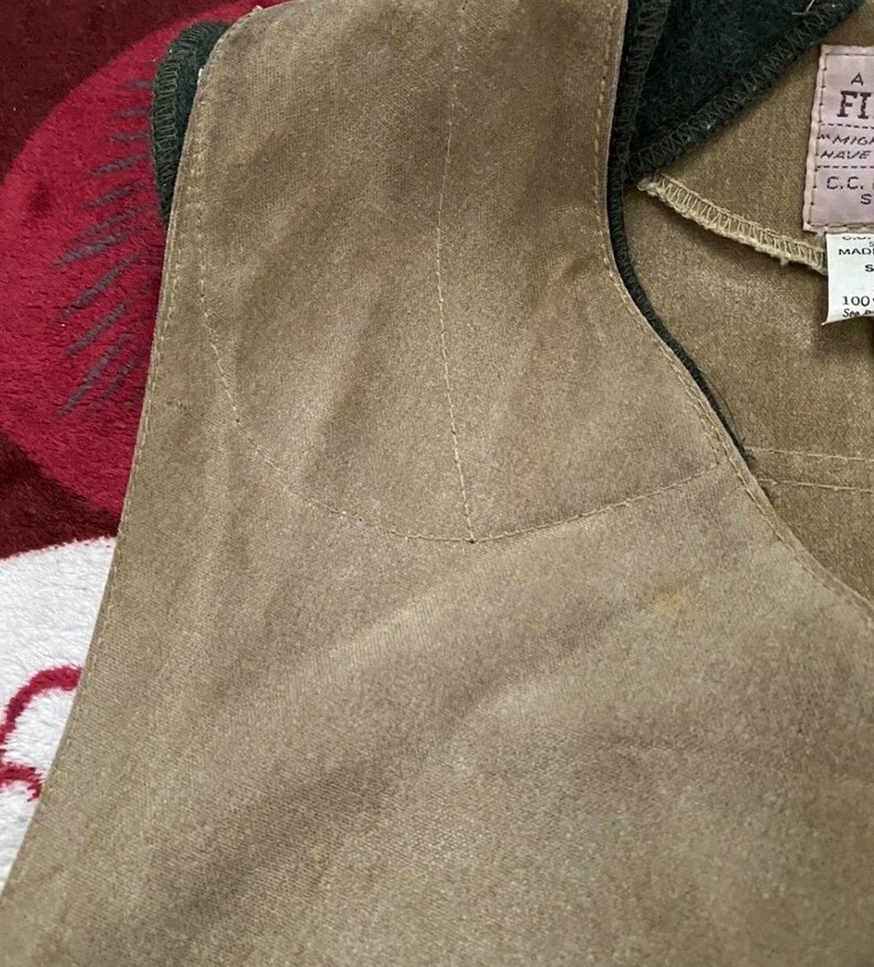 Peut inclure: Gros plan d'un gilet en daim beige avec un col vert fonc&eacute;. Le gilet a un d&eacute;tail de poche et des coutures visibles. Une &eacute;tiquette avec du texte est visible sur le c&ocirc;t&eacute; droit du gilet.