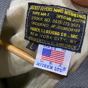 May include: Close-up of a vintage olive green bomber jacket label. The label features text including "JACKET FLYERS MANS INTERMEDIATE" and "AVIREX CLOTHING CO., INC.", with a small American flag. The jacket is size L.