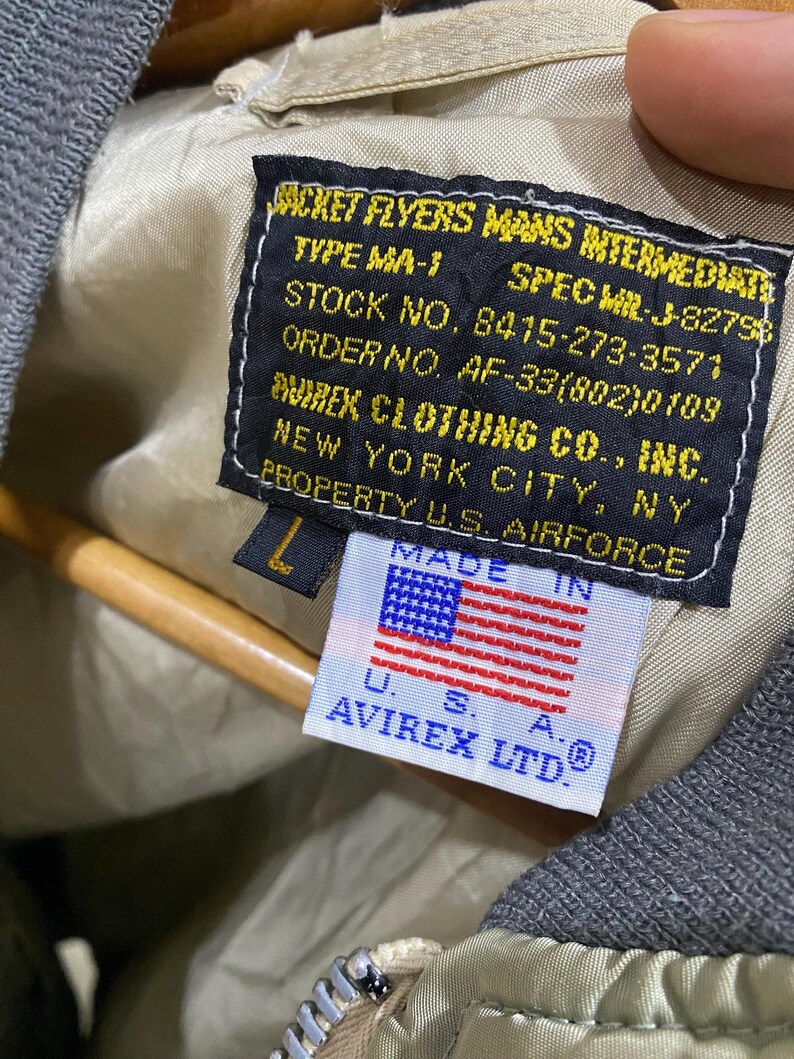 May include: Close-up of a vintage olive green bomber jacket label. The label features text including "JACKET FLYERS MANS INTERMEDIATE" and "AVIREX CLOTHING CO., INC.", with a small American flag. The jacket is size L.