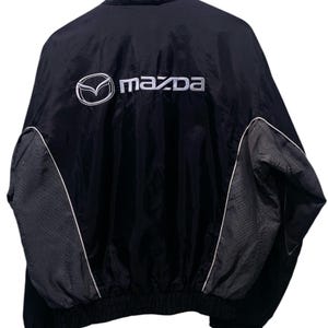 May include: Black and grey Mazda jacket with white trim and the Mazda logo embroidered on the back. The jacket has a high collar and a full zip. The sleeves and sides are grey, with white trim outlining the grey panels.