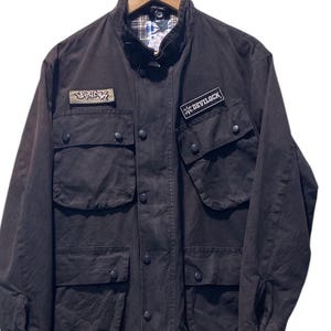 Puede incluir: Chaqueta marrón oscuro con múltiples bolsillos delanteros y cierres de botón. La chaqueta presenta dos parches, uno con el texto "DEVILOCK" y otro con texto estilizado. Cuelga de una percha de madera.