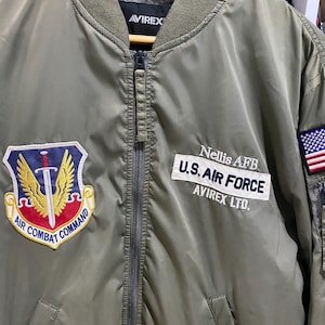 May include: An olive green bomber jacket featuring an Air Combat Command patch on the right chest and a U.S. Air Force Nellis AFB patch on the left chest. The left sleeve displays an American flag patch above a zippered utility pocket.