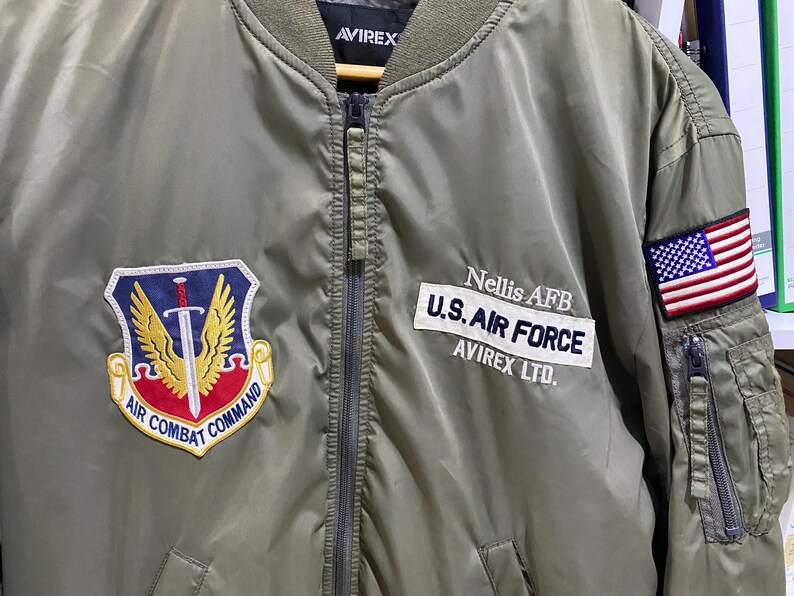May include: An olive green bomber jacket featuring an Air Combat Command patch on the right chest and a U.S. Air Force Nellis AFB patch on the left chest. The left sleeve displays an American flag patch above a zippered utility pocket.