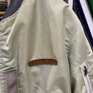 May include: A light green bomber jacket with a brown leather patch that reads "AVIREX LTD." The jacket has a gray collar and a zipper on the sleeve. The jacket is made of a smooth, shiny material.