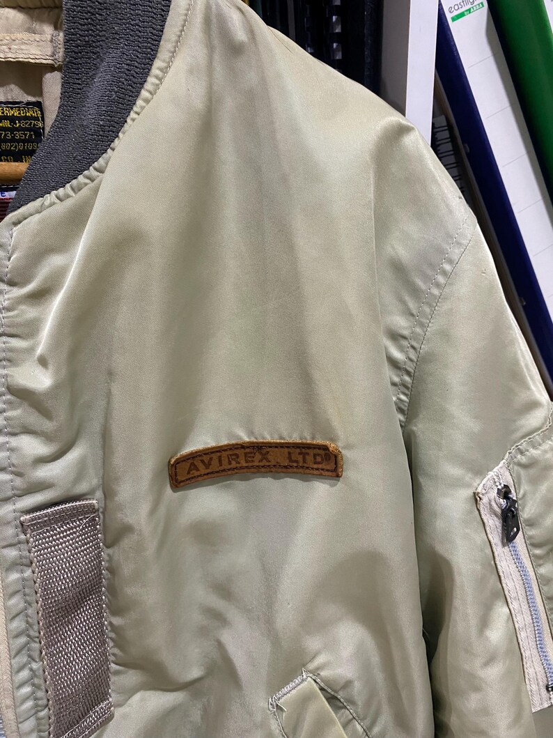 May include: A light green bomber jacket with a brown leather patch that reads "AVIREX LTD." The jacket has a gray collar and a zipper on the sleeve. The jacket is made of a smooth, shiny material.