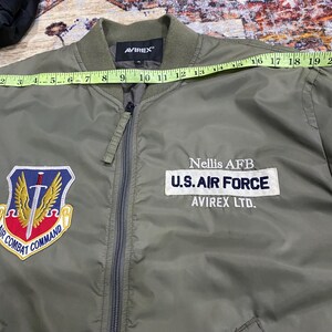 May include: An olive green bomber jacket with a front zipper, featuring an Air Combat Command patch, a U.S. Air Force Nellis AFB patch, and a U.S. flag patch on the sleeve. A yellow measuring tape across the chest shows a width of 22 inches.