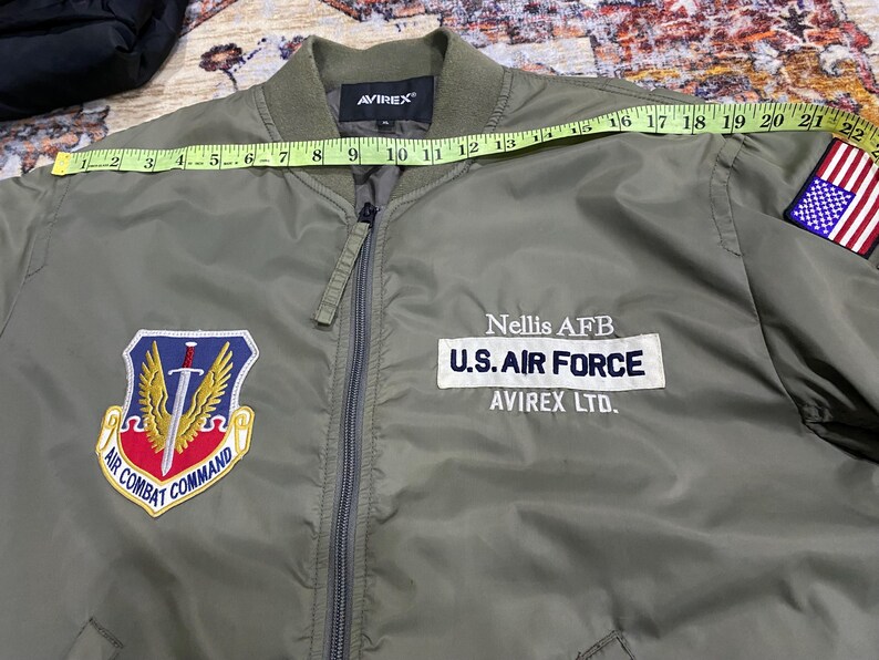 May include: An olive green bomber jacket with a front zipper, featuring an Air Combat Command patch, a U.S. Air Force Nellis AFB patch, and a U.S. flag patch on the sleeve. A yellow measuring tape across the chest shows a width of 22 inches.