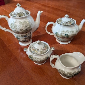 May include: A vintage tea set comprising two teapots, a sugar bowl, and a creamer. The white porcelain pieces feature a landscape design and floral detailing. The teapots have curved spouts and handles, typical of the era.