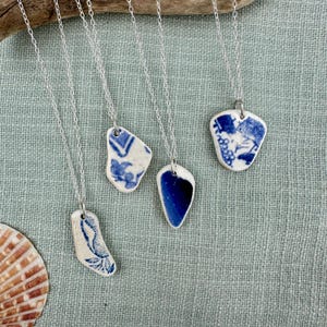 May include: Four silver necklaces with unique pendants. Each pendant is a piece of white ceramic with blue patterns or solid blue. The necklaces are displayed on a light blue fabric, with a seashell in the corner.