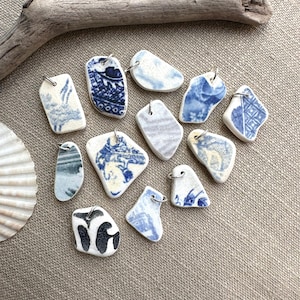 May include: A selection of ceramic shard pendants in various shapes, adorned with blue and white patterns. Each pendant has a silver-tone metal loop for suspension. The pendants are displayed on a neutral fabric, with a piece of driftwood visible in the background.