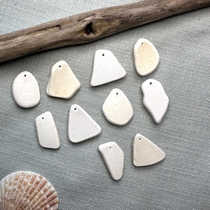 May include: A collection of off-white, irregularly shaped pendants with small holes for stringing. Some pendants have a slightly aged appearance. A piece of driftwood rests above the pendants on a textured, light blue surface. A seashell is in the lower left corner.