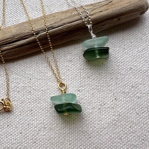 Könnte beinhalten: Nahaufnahme von drei Halsketten mit Anhängern aus Meerglas. Eine Halskette hat eine goldene Kette, der Anhänger besteht aus zwei grünen Meerglasstücken. Eine andere Halskette hat eine silberne Kette mit einem ähnlichen Anhänger. Der Hintergrund ist eine strukturierte, cremefarbene Oberfläche.