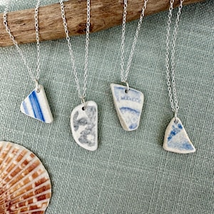 May include: Four silver chain necklaces, each with a different, irregularly shaped pendant. The pendants are white with blue and grey patterns, resembling pottery shards. A seashell is visible in the lower left corner.