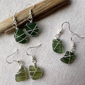 May include: Four pairs of handmade earrings with green sea glass. Each piece of sea glass is wrapped in silver-coloured wire and suspended from silver-coloured fish hook ear wires. The earrings are displayed on a light-coloured fabric, with a piece of driftwood.