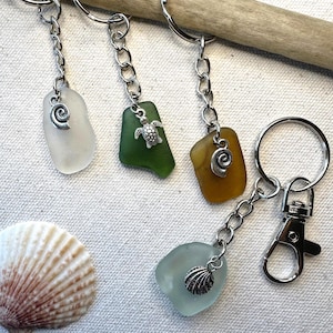 May include: Keychains featuring sea glass in various colors, including white, green, and amber. Each keychain has a silver-toned charm, such as a spiral, a turtle, or a seashell. The keychains are attached to silver chains and key rings.