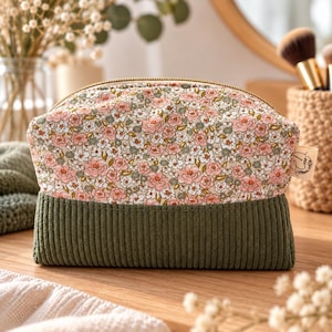 Peut inclure: Une trousse zippée composée d'une partie supérieure à motif floral avec des fleurs roses et blanches sur fond beige, associée à une partie inférieure en velours côtelé vert olive. Une petite étiquette en tissu avec un logo est fixée sur le côté.