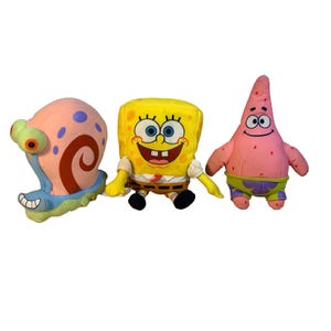 May include: Three plush toys of characters from Spongebob Squarepants. The toys include a yellow Spongebob, a pink starfish named Patrick, and a pink snail named Gary. The toys are made of soft fabric and have embroidered details.