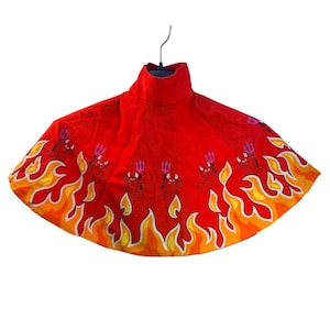 May include: A bright red circular cape featuring a repeating pattern of small cartoon devils holding purple tridents. The bottom hem is decorated with a vibrant orange and yellow flame design that wraps around the entire lower edge of the garment.