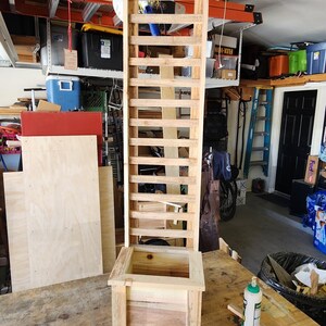 Puede incluir: Una jardinera alta de madera clara con un enrejado vertical integrado. La estructura cuenta con una base cuadrada con cuatro patas cortas y un panel trasero de rejilla rectangular que se extiende hacia arriba, hecho de listones de madera natural.