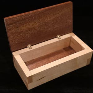 May include: A rectangular wooden box with a hinged lid, partially open. The box has a light-coloured body and a darker brown lid. The hinges are visible. The box is likely for storage or display, with a simple, elegant design.