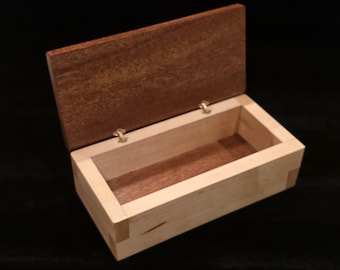 May include: Open wooden box with a hinged lid. The box is rectangular with a light-colored wood body and a darker brown lid. The lid is slightly ajar, revealing the interior. The box is likely used for storage or display.