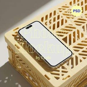 May include: A smartphone with a blank white screen and a black frame resting on a pale yellow plastic storage crate with a lattice design. The screen displays the text Phone Mockup by Lynie and a yellow badge with the text PSD in blue letters.