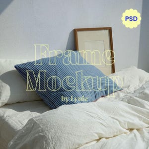 May include: A blue and white striped pillow rests on a bed with white linens. A brown framed picture leans against the wall. The image has the text "Frame Mockup by Lynie" and a yellow "PSD" graphic.