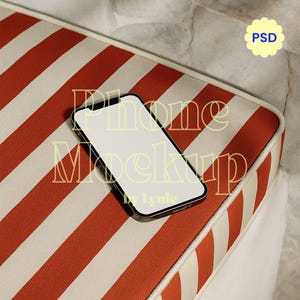 Peut inclure: Un smartphone à écran blanc vierge est posé sur un coussin rayé. Le coussin présente des rayures rouges et crème alternées. L'arrière-plan comprend un mur en pierre et un graphisme jaune en forme de fleur avec les lettres "PSD". Le texte "Phone Mockup by Lyuie" est superposé à l'image.