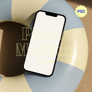 May include: A black smartphone with a blank white screen, positioned on a blue, tan, and brown inflatable ring. The words "Phone Mockup" are displayed in a light yellow font. A small, light blue flower-shaped sticker with the letters "PSD" is in the upper right corner.