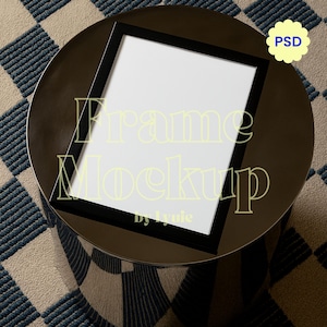 May include: A black picture frame mockup with a white interior, resting on a dark brown, circular table. The table is set on a patterned rug with a blue and beige checkered design. The text "Frame Mockup" is visible on the frame.