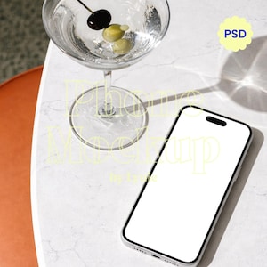 Peut inclure: Un verre à martini avec un liquide clair, une olive noire et deux olives vertes est posé à côté d'un smartphone argenté avec un écran blanc vierge. Les éléments sont sur une surface en marbre blanc. Un graphique "PSD" jaune est dans le coin supérieur droit.