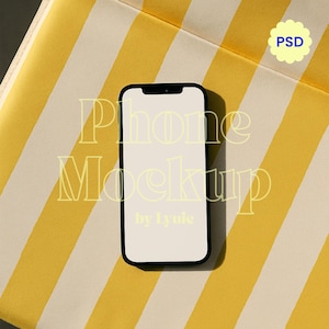 May include: A black smartphone mockup with a blank white screen rests on a yellow and white striped surface. The text "Phone Mockup by lyuie" is displayed on the phone. A small "PSD" icon is in the upper right corner.