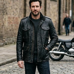 Pode incluir: Casaco de couro preto com vários bolsos, gola e fecho de correr. O casaco é usado com um suéter cinza escuro e jeans azuis. Uma motocicleta está estacionada ao fundo.