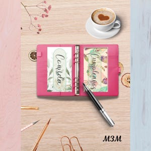 May include: A pink binder with the words "Comida" and "Cumpleaños" on inserts. A black pen rests on the open binder. A cup of coffee with a heart-shaped design is in the background. The text "M3M" is at the bottom.